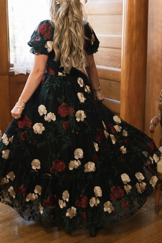 A woman with long wavy blonde hair stands indoors by wooden paneling, holding out the full skirt of the Ivy City Co Amora Dress in Obsidian Rose Embroidery—a black tulle ball gown beautifully adorned with red and white floral patterns.