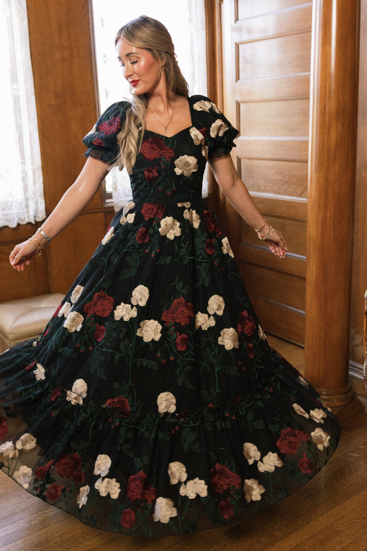 In a graceful twirl, a woman stands indoors on a wooden floor near a wooden wall and lace-curtained window, wearing the Ivy City Co Amora Dress in Obsidian Rose Embroidery—a flowing black dress adorned with red and white floral patterns and featuring puff sleeves with a sweetheart neckline.