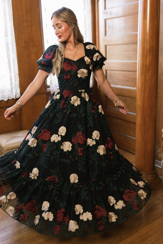 In a graceful twirl, a woman stands indoors on a wooden floor near a wooden wall and lace-curtained window, wearing the Ivy City Co Amora Dress in Obsidian Rose Embroidery—a flowing black dress adorned with red and white floral patterns and featuring puff sleeves with a sweetheart neckline.
