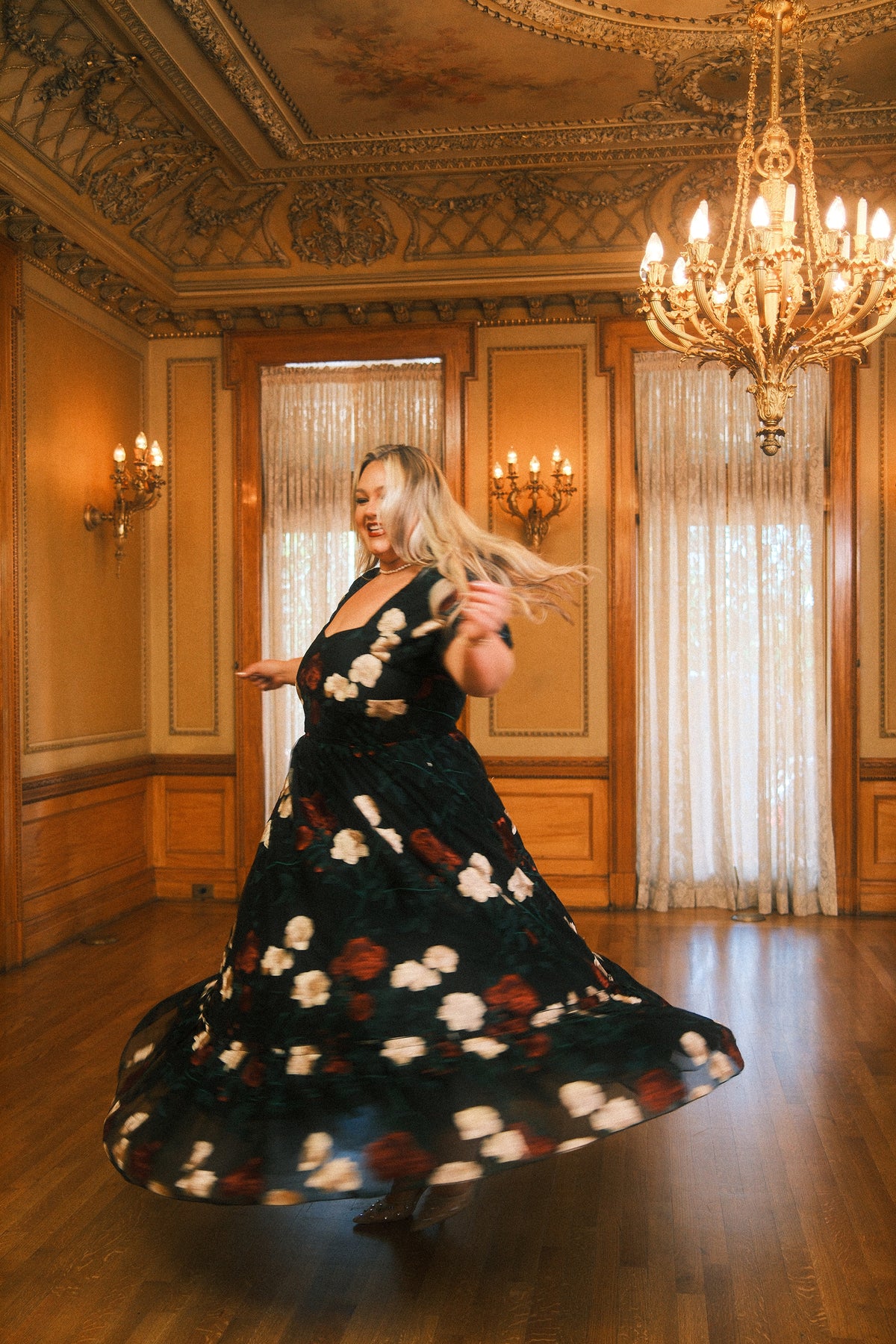 A woman wearing the Amora Dress in Obsidian Rose Embroidery by Ivy City Co, featuring a puff sleeve and sweetheart neckline, twirls gracefully in a grand, ornately decorated room with chandeliers, wooden floors, and tall windows draped with sheer curtains.