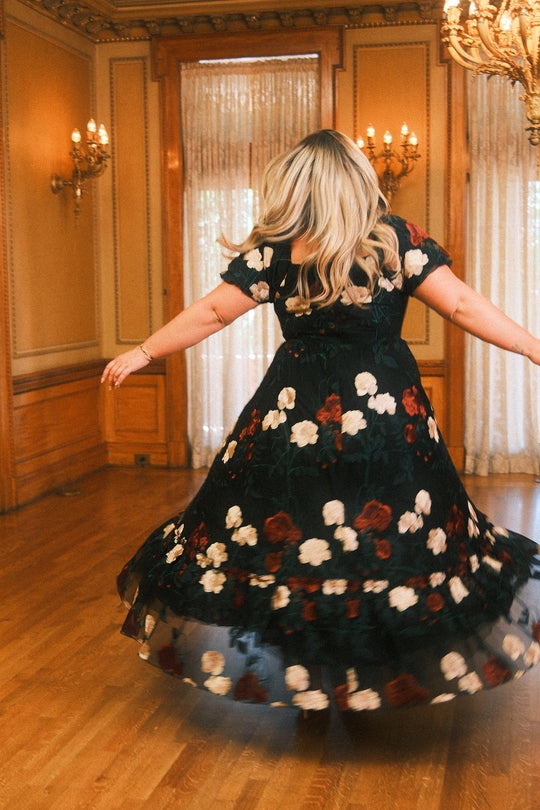 A woman with long blonde hair twirls in the Ivy City Co Amora Dress in Obsidian Rose Embroidery—a black, floral, puff sleeve dress with a sweetheart neckline—in an elegant, wood-paneled room illuminated by chandeliers and tall windows draped with sheer curtains.