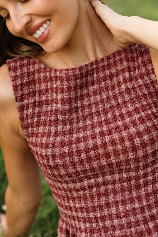 A woman smiling, wearing the sleeveless Ana Dress in Cranberry Popcorn Plaid by Ivy City Co, featuring a textured maroon and pink checked pattern with a smocked bodice, touches her neck with one hand while standing outdoors on green grass. This stylish dress is also maternity friendly.