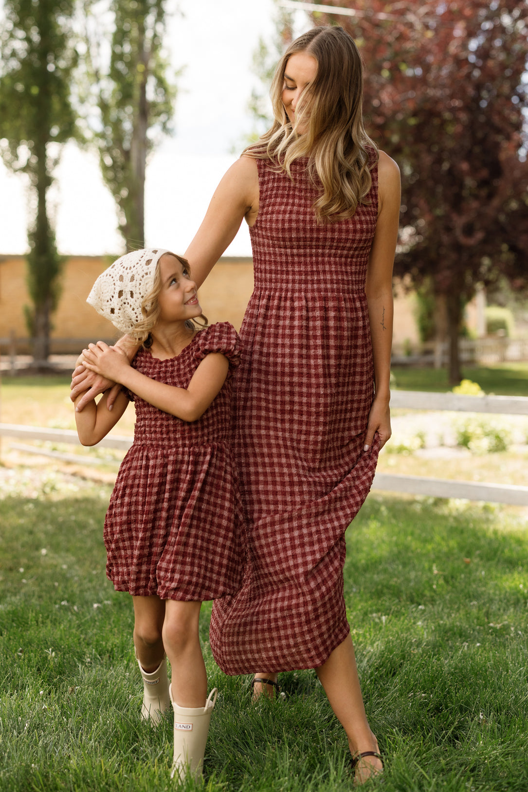 A woman and a young girl, both wearing matching Ivy City Co Ana Dresses in Cranberry Popcorn Plaid with smocked bodices, smile and hold hands while walking on grass outdoors. The girl pairs her dress with a white knit hat and boots, highlighting the maternity-friendly design that ensures comfort for all ages.