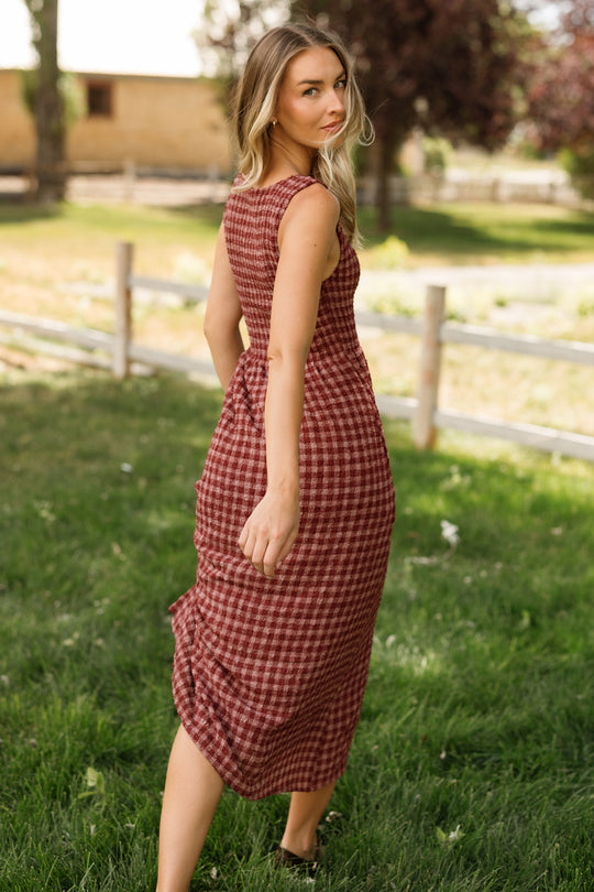 A woman walks outdoors on grass, looking back over her shoulder, wearing the Ivy City Co Ana Dress in Cranberry Popcorn Plaid—a sleeveless style with a smocked bodice. This maternity-friendly dress offers comfort and ease. A wooden fence and trees appear in the background.