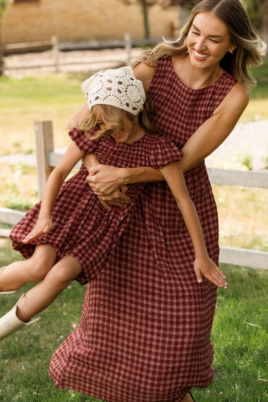 An adult wearing the Ivy City Co Ana Dress in Cranberry Popcorn Plaid—with its sleeveless cut and smocked bodice—spins a smiling child dressed in a matching outfit and white crochet hat outside on the grass near a wooden fence. This style is maternity friendly and ideal for playful moments together.