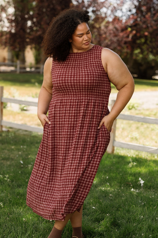 A woman with curly hair stands on grass near a wooden fence and trees, wearing the Ana Dress in Cranberry Popcorn Plaid by Ivy City Co—a sleeveless dress featuring a smocked bodice, practical pockets, and a charming red plaid pattern. This stylish piece is maternity friendly, offering both comfort and elegance.