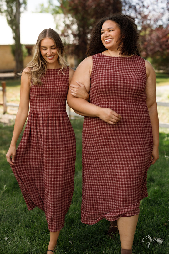 Two women walk arm in arm outdoors, both smiling and enjoying a sunny day as they wear sleeveless, ankle-length Ana Dresses in Cranberry Popcorn Plaid by Ivy City Co. The smocked bodice and flowy design of the dresses make them comfortable and maternity friendly. Lush grass and trees form the backdrop to their cheerful outing.