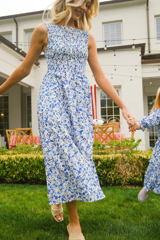 A woman smiles and walks barefoot on grass, holding a child’s hand, wearing the Ivy City Co Ana Dress in Ditsy Cobalt Floral in front of a house with large windows and outdoor string lights.