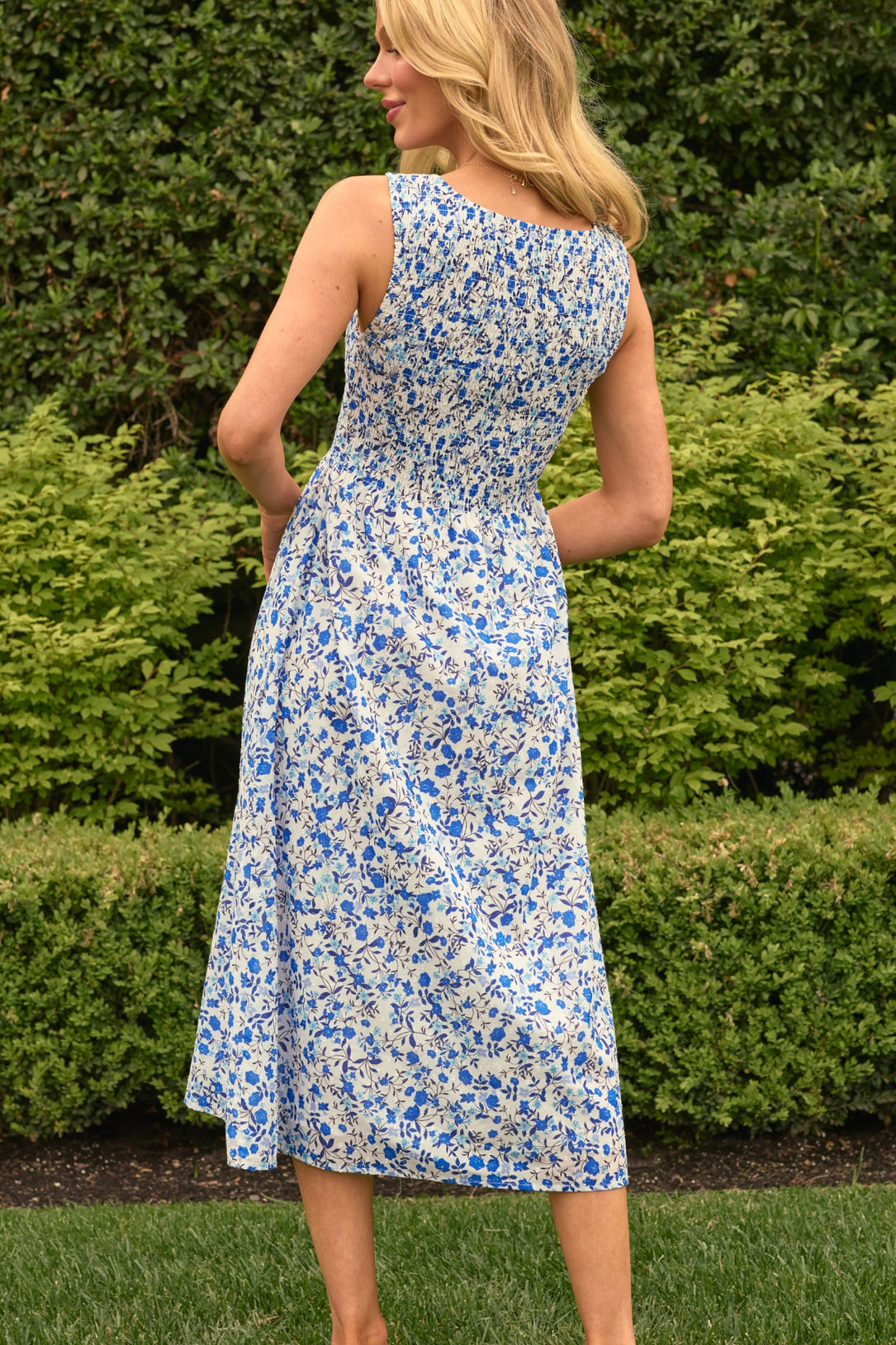 A woman with long blonde hair stands outdoors facing away, wearing the Ana Dress in Ditsy Cobalt Floral by Ivy City Co. Green bushes and grass are visible in the background.