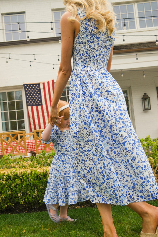 A woman and a young girl wear matching Ivy City Co Ana Dresses in Ditsy Cobalt Floral as they hold hands and walk in a garden, with an American flag hanging on a white house in the background.