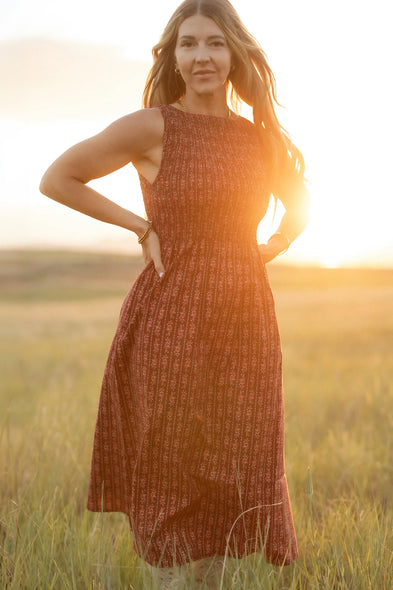 A woman stands in a sunlit field wearing the Ivy City Co Ana Dress in Sequoia Felicity Floral, looking at the camera with her hands on her hips against a softly blurred, warm evening background.