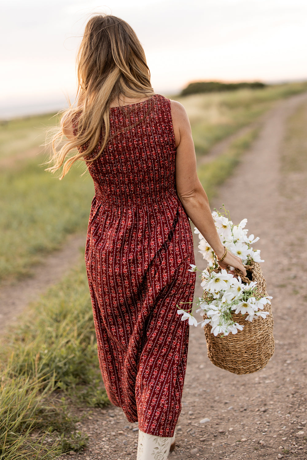 A woman with long hair wearing the Ivy City Co Ana Dress in Sequoia Felicity Floral walks along a dirt path through a grassy field, holding a woven basket filled with white flowers.