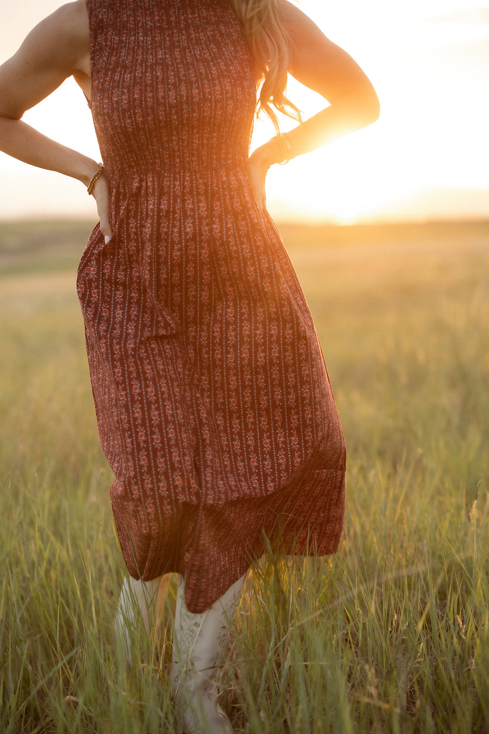 A woman wearing the Ivy City Co Ana Dress in Sequoia Felicity Floral stands hands-on-hips in a sunlit grassy field at sunset; her head is out of frame and golden light bathes the scene.