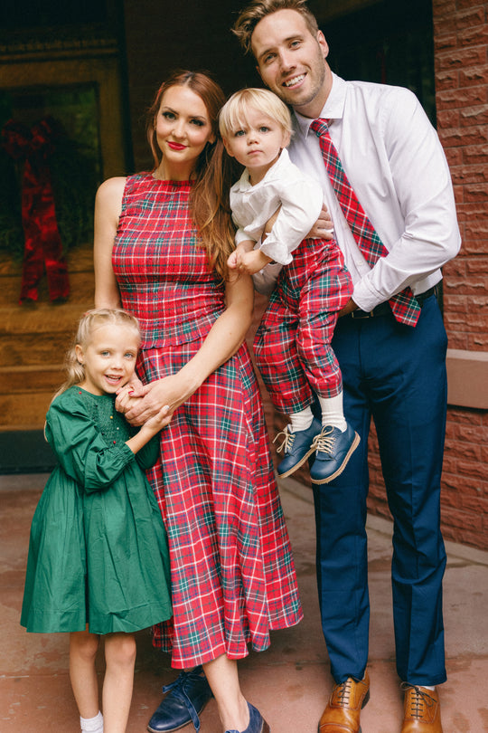 A family of four poses outside, smiling and standing close together. The mother looks festive in the Ivy City Co Ana Skirt in Christmas Cheer Plaid, which matches her son’s red plaid outfit. The father complements them with a white shirt and coordinating red plaid tie, while the daughter wears a green dress.