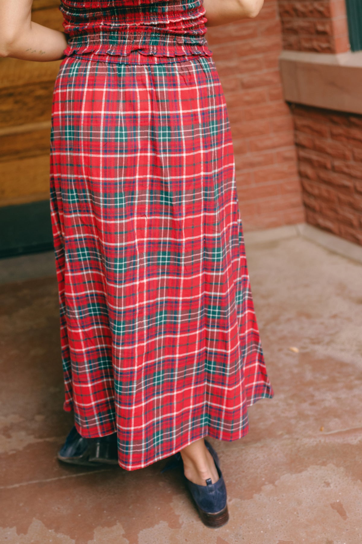 A person wearing the Ivy City Co Ana Skirt in Christmas Cheer Plaid and black shoes stands on a concrete surface near a brick wall, their upper body mostly out of frame. The classic skirt brings a festive touch to the simple scene.