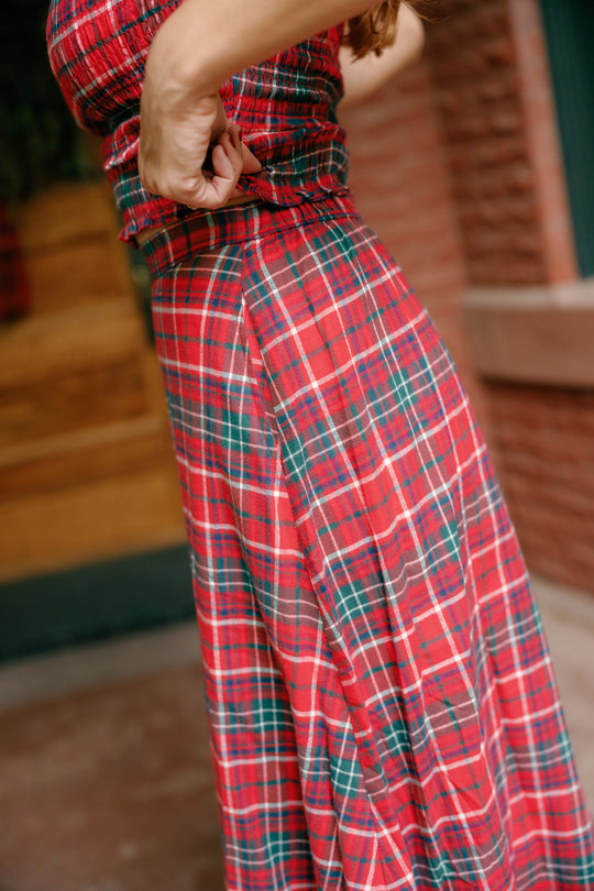 Standing outdoors near a brick wall and wooden door, a person adjusts the Ana Skirt in Christmas Cheer Plaid by Ivy City Co—their hands highlighting the festive red and green patterned cotton A-line fabric.