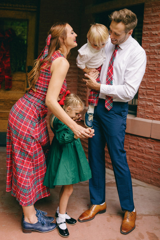 A smiling family of four poses together outside in festive outfits: the mother wears a classic holiday skirt, while the daughter is dressed in Ivy City Co’s Ana Skirt in Christmas Cheer Plaid paired with a green dress. The father and son complement them in crisp white shirts and dress shoes.