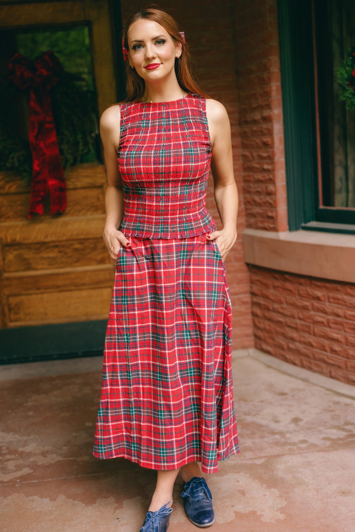 A woman stands outside, smiling with her long auburn hair cascading over her shoulders. She wears the Ivy City Co Ana Skirt in Christmas Cheer Plaid—a sleeveless A-line cotton skirt featuring a vibrant red plaid pattern and convenient pockets—paired with blue shoes. A brick wall and wooden door provide the backdrop.