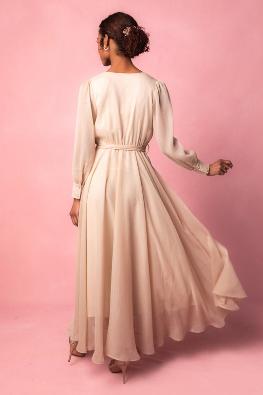 A woman stands against a pink background, turned slightly away, wearing the Ivy City Co Andie Dress in Champagne—a long, flowing maxi style. Her hair is styled in a bun adorned with decorative pins, and she completes the look with nude heels.