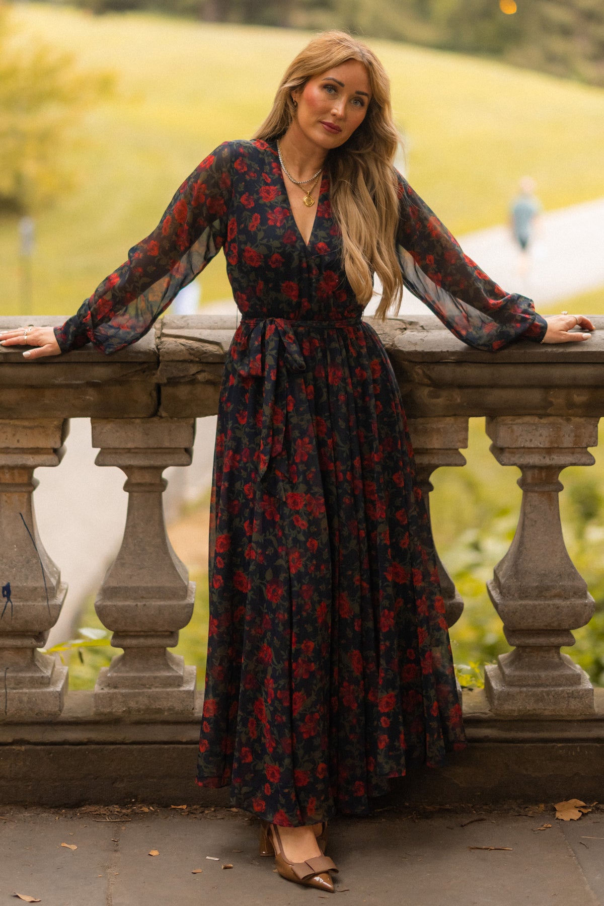 A woman with long, wavy hair and layered necklaces stands outdoors in a sunlit, grassy park, leaning on a stone railing. She is wearing the Ivy City Co Andie Dress in Scarlet Blooms—a long fall floral maxi dress with sheer sleeves.