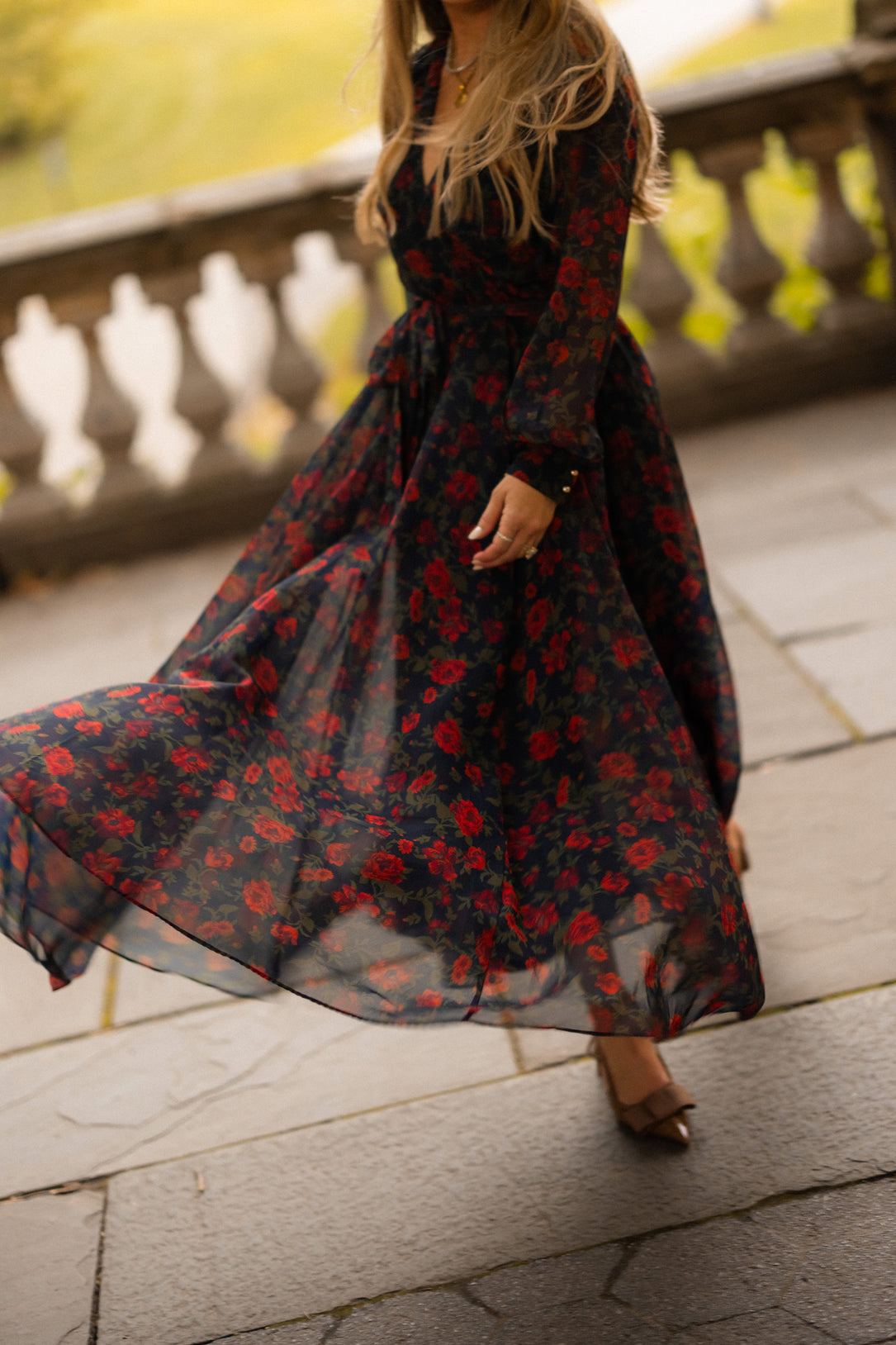 A woman twirls on a stone patio in the Ivy City Co Andie Dress in Scarlet Blooms, her face out of frame, with blurred greenery and a stone railing in the background.
