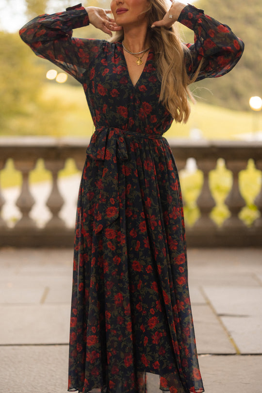Wearing the Ivy City Co Andie Dress in Scarlet Blooms, a woman stands outdoors adjusting her hair. Her face is partially out of frame, with greenery and a stone balustrade in the background.