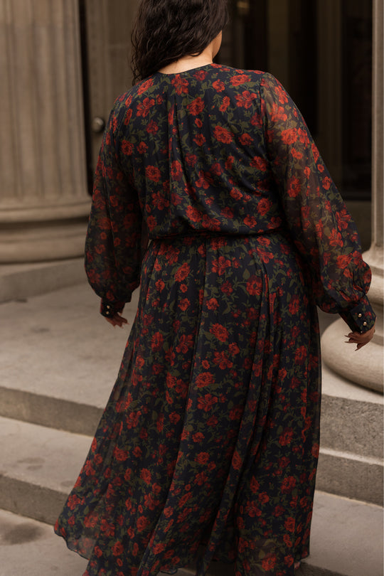 A person with wavy dark hair walks up stone steps, wearing Ivy City Co’s Andie Dress in Scarlet Blooms—a long, sheer black maxi dress featuring red floral patterns and long sleeves. The background shows tall columns and part of a building.