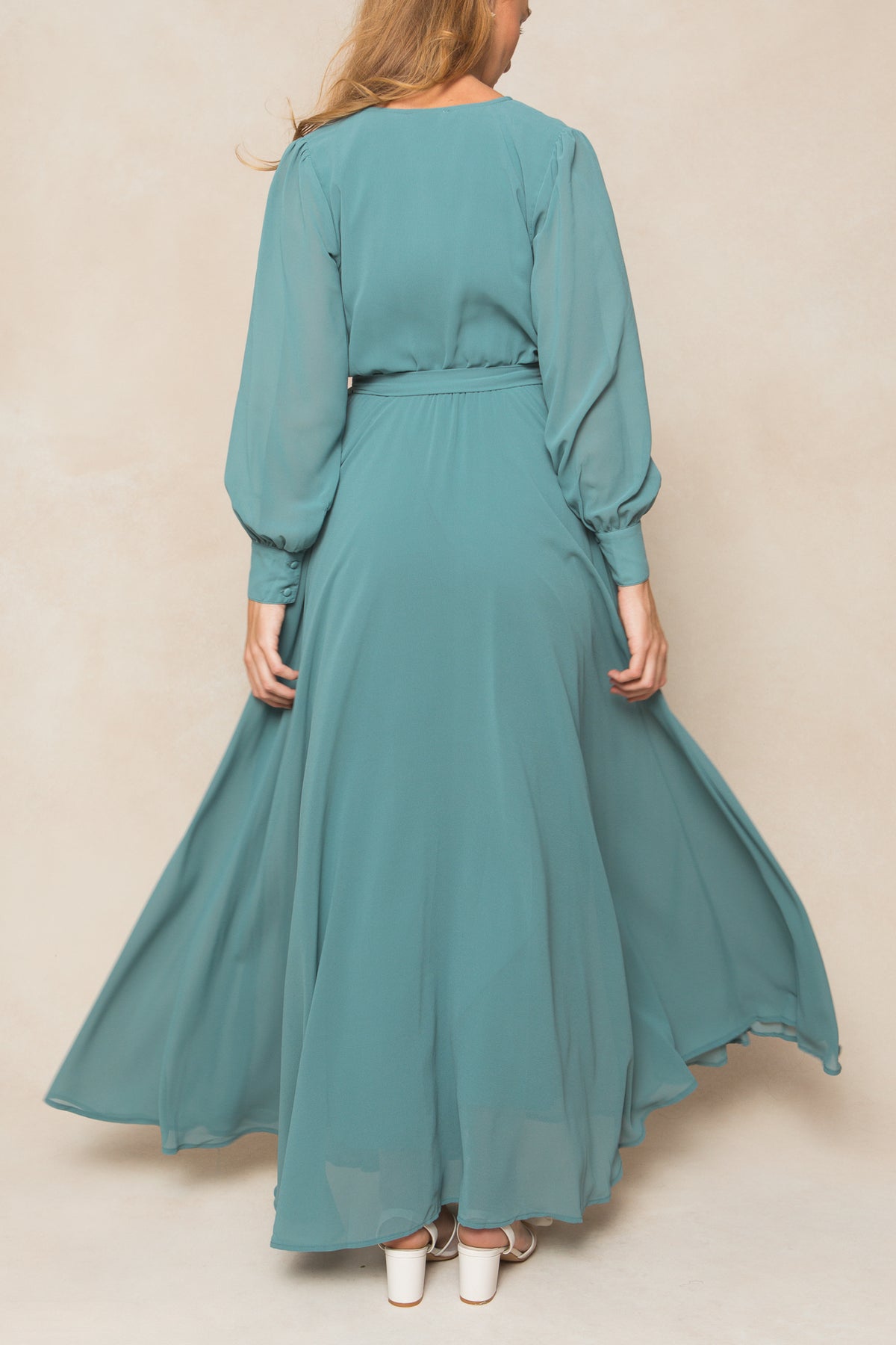 A woman with long hair stands with her back to the camera in the Ivy City Co Andie Dress in Seaglass Chiffon, featuring long sleeves and a belted waist. She pairs it with white heels against a neutral background—ideal for fall bridesmaids.