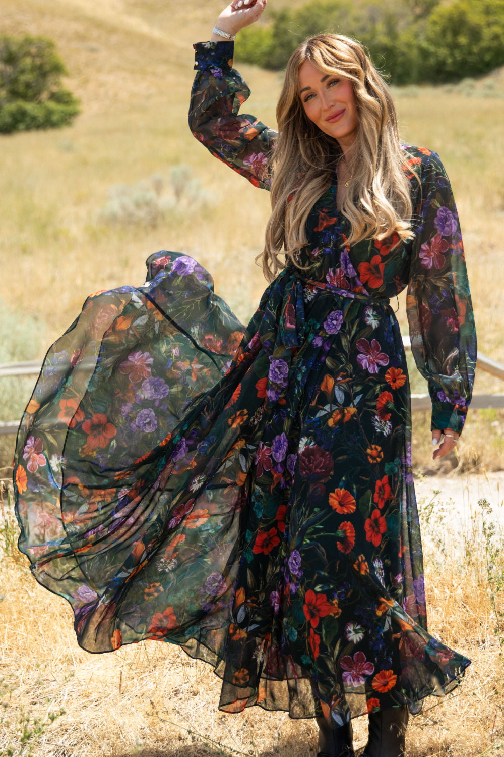 A woman stands outdoors in the Ivy City Co Andie Dress in Evergreen Vivid Meadow—a flowing, sheer, long-sleeved black maxi dress adorned with colorful floral prints. Smiling, she lifts her nursing-friendly chiffon dress slightly, surrounded by dry grass and greenery.