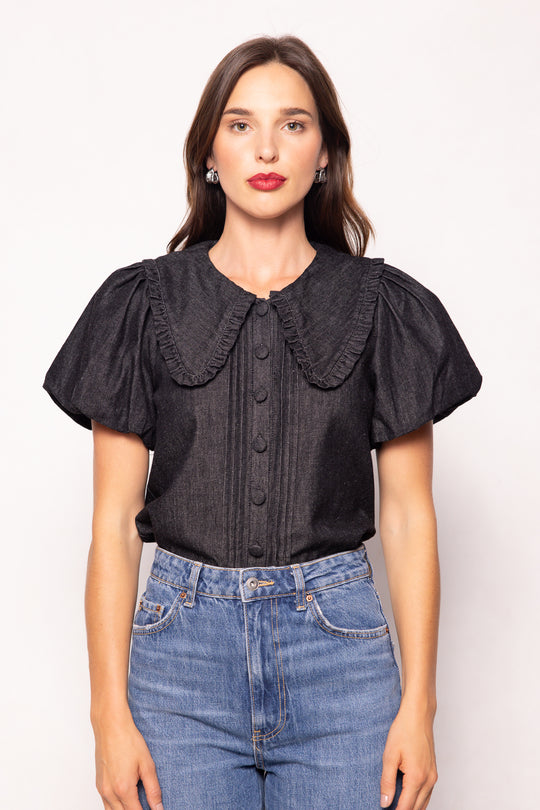 Woman wearing a black blouse with ruffled collar and short sleeves, standing against a white background.