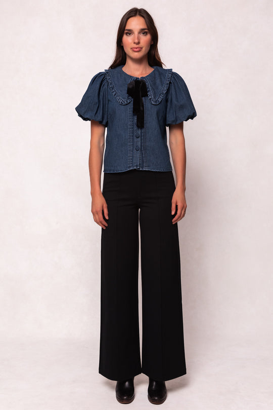 A woman faces forward in the Ivy City Co Andrea Blouse in Denim with Velvet Tie, featuring a large ruffled collar, paired with high-waisted, wide-leg black trousers and black shoes against a plain light background.