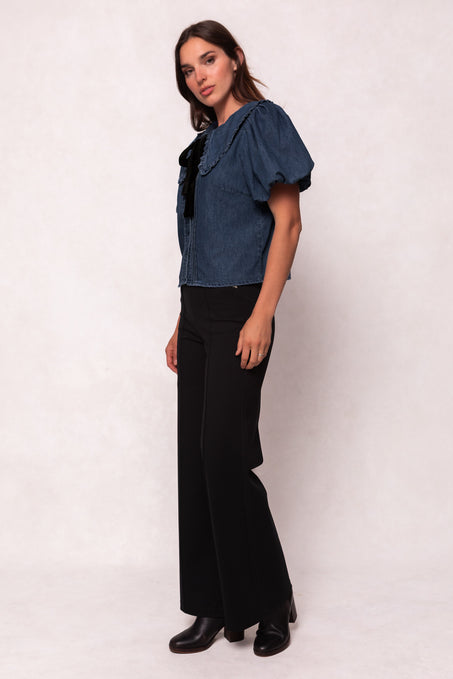 A woman stands against a light background wearing the Ivy City Co Andrea Blouse in Denim with Velvet Tie, paired with black wide-leg pants and black ankle boots. She looks at the camera with a relaxed expression.