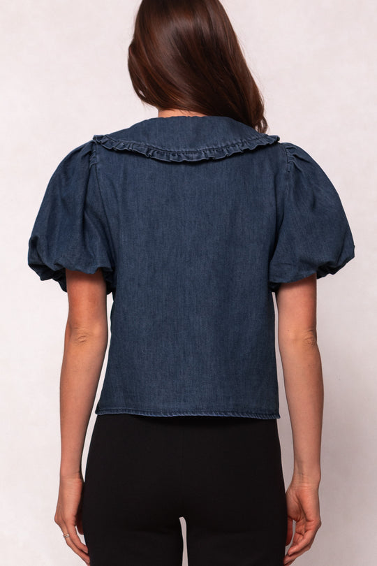 A woman wears the Andrea Blouse in Denim with Velvet Tie by Ivy City Co, featuring a ruffled collar for an elegant, nursing-friendly style.