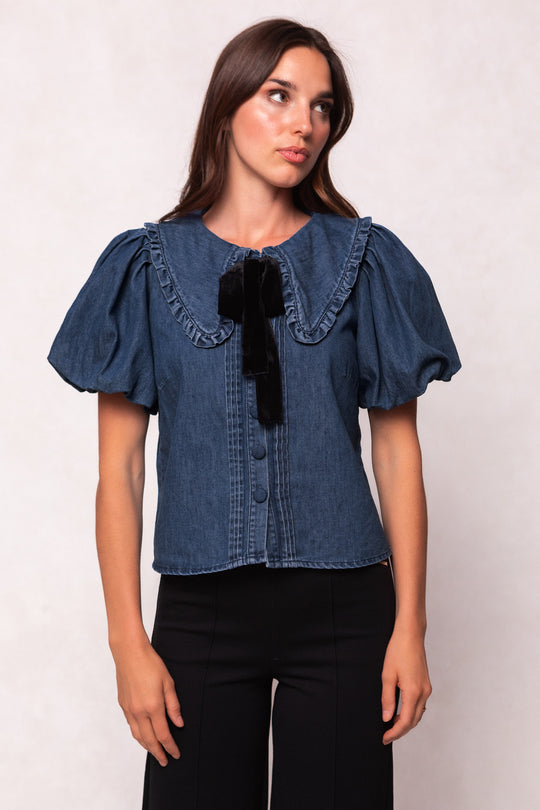 A woman wears the Ivy City Co Andrea Blouse in Denim with a velvet tie, featuring puffed sleeves and a ruffled collar, paired with black pants against a plain light background.