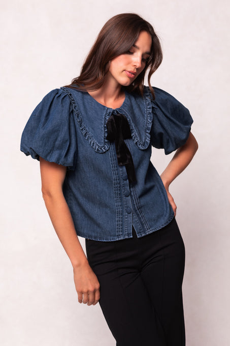 A woman with long brown hair wears the Ivy City Co Andrea Blouse in Denim with Velvet Tie, featuring puffed sleeves, a ruffled collar, and a black bow, paired with black pants against a light background. Her head is slightly tilted down.