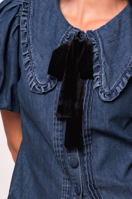 A close-up shows part of the neck, shoulders, and upper chest of a person wearing the Ivy City Co Andrea Blouse in Denim with Velvet Tie, featuring ruffled edges and a large black velvet bow at the collar.