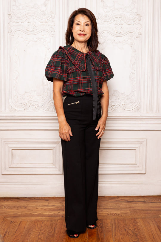 Woman wearing a plaid top and black pants against a white wall.