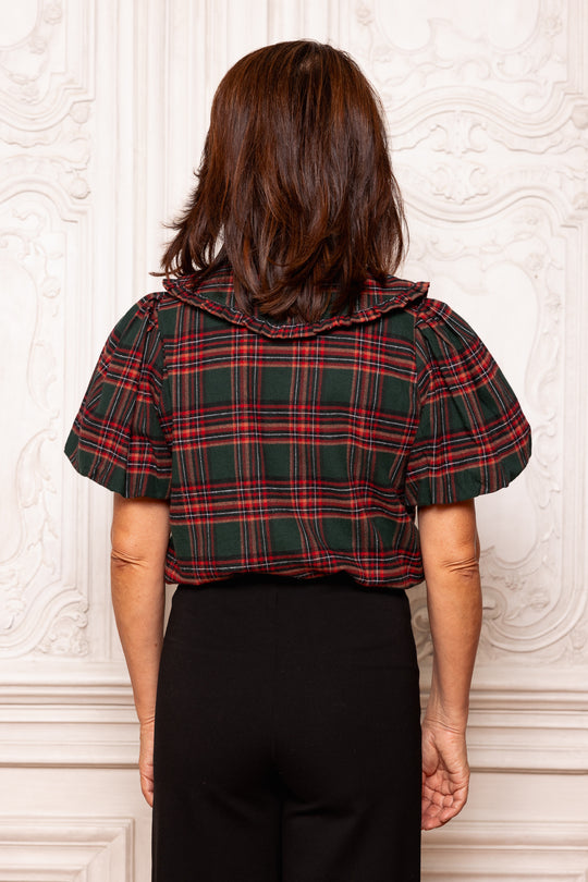 Person wearing a plaid shirt with ruffled shoulders against a decorative wall.