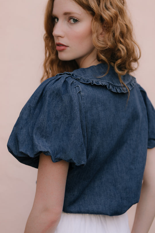 A woman with wavy red hair looks over her shoulder, wearing the Ivy City Co Andrea Blouse in Denim with Velvet Tie and a white skirt, set against a light pink background.