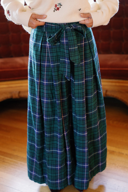 A person wearing the Angela Skirt in Emerald Plaid by Ivy City Co, styled with a white sweater adorned with small embroidered flowers, stands on a wooden floor in front of a brown couch—perfect for charming holiday portraits.