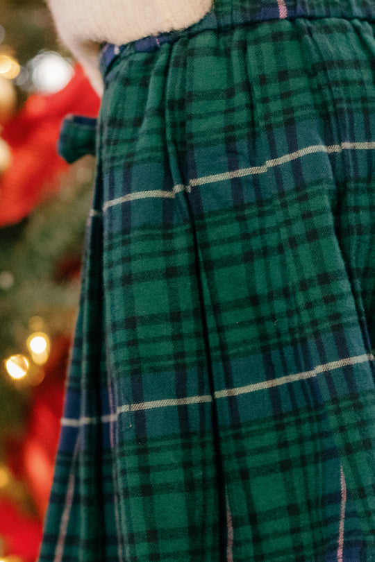 Close-up of the Angela Skirt in Emerald Plaid by Ivy City Co, showcasing its green and blue pleated design—perfect for holiday portraits—worn near a decorated Christmas tree with blurred lights and red ribbon in the background.
