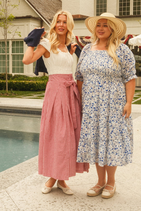 Two women stand poolside outdoors. One wears Ivy City Co's Angela Skirt in Red Gingham with a white blouse, the other a blue floral dress and wide-brimmed hat. Both smile, showcasing chic summer style against greenery and a house backdrop.