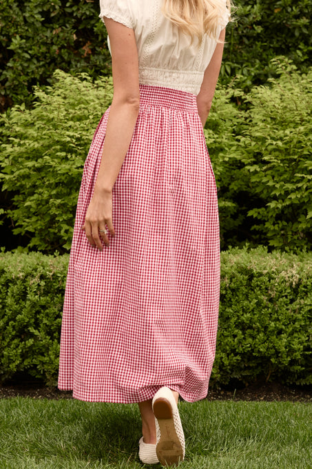 A woman stands outdoors with her back to the camera, wearing Ivy City Co's Angela Skirt in Red Gingham—a summer essential. Her long blonde hair flows as lush greenery and hedges fill the background.