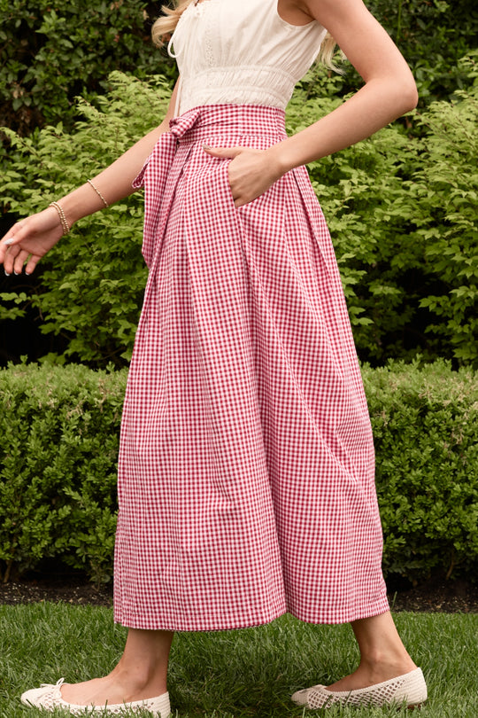 A woman walks on grass before green hedges wearing the Ivy City Co Angela Skirt in Red Gingham with a bow and a sleeveless white top. Only the lower half of her face to her feet are visible, creating a perfect summer look.