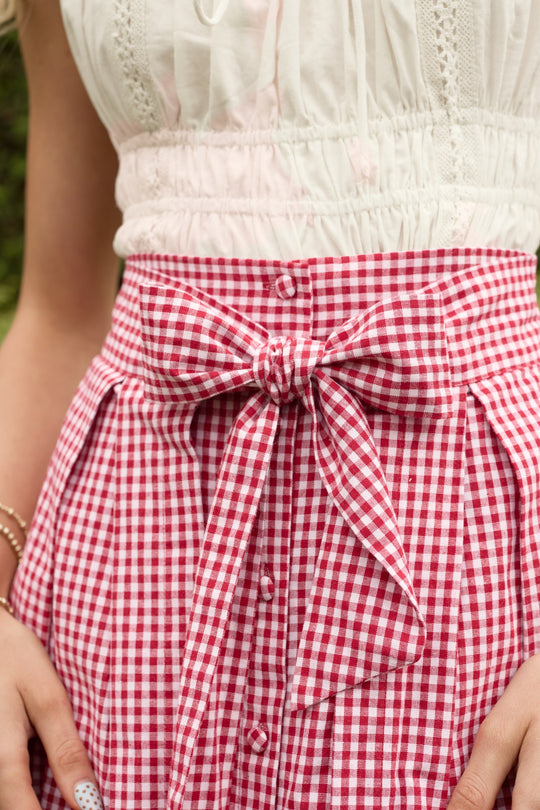 A person wears the Ivy City Co Angela Skirt in Red Gingham—a mini skirt featuring front buttons and a large waist bow—paired with a white sleeveless top, making it a charming summer wardrobe addition. Their hands rest at their sides.