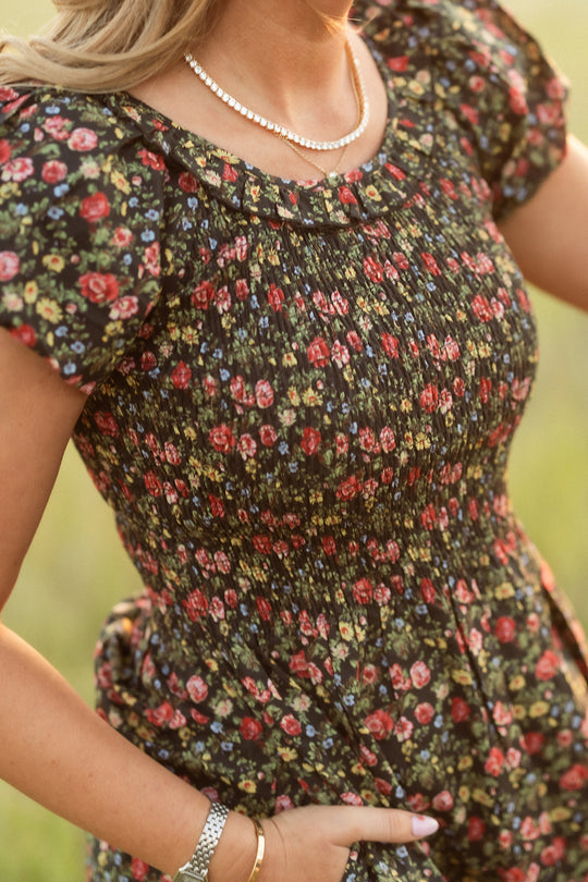 A woman in the Ivy City Co Annalise Dress in Obsidian Vintage Poppies, paired with a pearl necklace, silver watch, and gold bracelets, stands outdoors with her hand in her pocket. The image is softly focused and warmly lit.