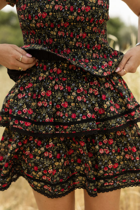 A person holds the Ivy City Co Annalise Skirt in Obsidian Vintage Poppies—black with red and yellow floral print and lace trim—outdoors, standing before a blurred grassy field.