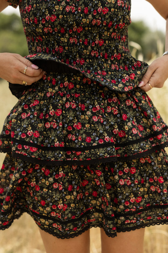 A person holds the Ivy City Co Annalise Skirt in Obsidian Vintage Poppies—black with red and yellow floral print and lace trim—outdoors, standing before a blurred grassy field.