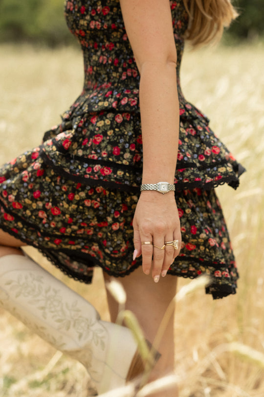A woman stands in a grassy field wearing the Ivy City Co Annalise Skirt in Obsidian Vintage Poppies and white embroidered boots. Her left arm, decorated with rings and a watch, hangs relaxed by her side. Her face is not visible.