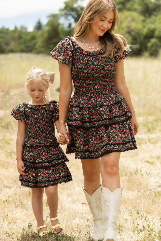 A woman and young girl wear matching Ivy City Co Annalise Skirts in Obsidian Vintage Poppies while holding hands in a grassy field. The woman pairs hers with white cowboy boots; the girl sports yellow sandals and a ponytail.